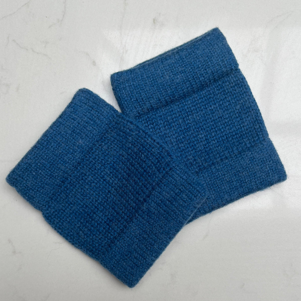 Two teal blue coloured cashmere knit wristwarmers cuffs with ribbed elastic edges, displayed on a light gray marble surface.