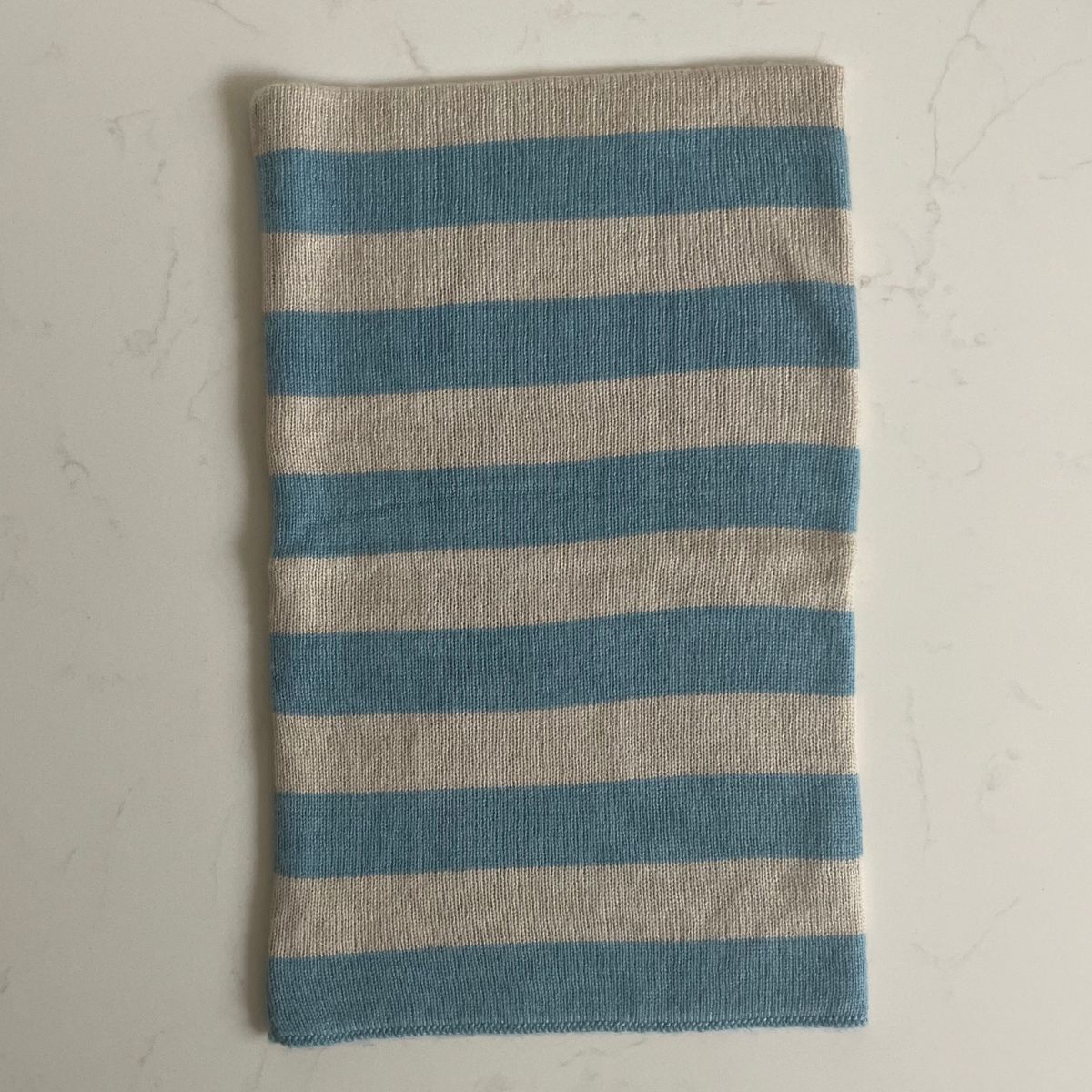 A striped blue and beige cashmere snood laid out on a light gray background
