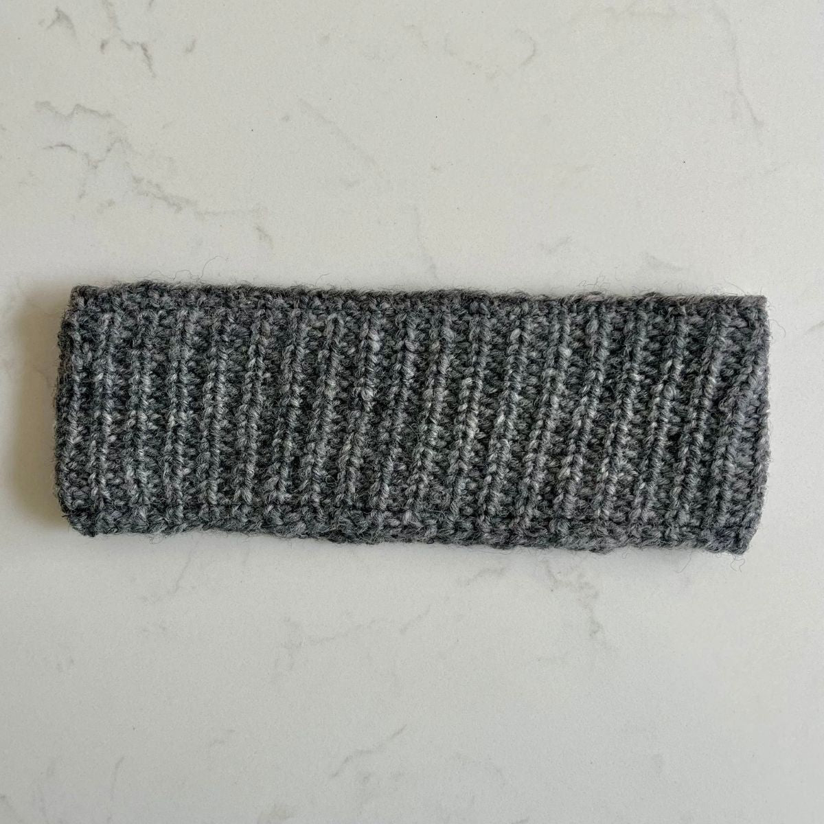 Gray plain knitted woollen headband with a black fleece inner lining on a light gray marble background