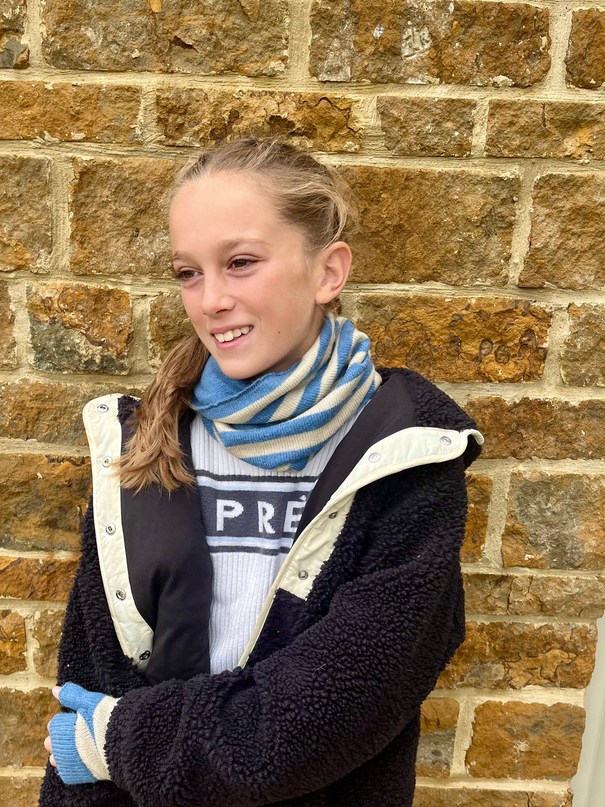 A young girl smiling in front of a rustic brick wall, wearing a blue and beige striped cashmere snood, a white sweater with blue stripes, and blue & beige knit wristwarmer gloves.