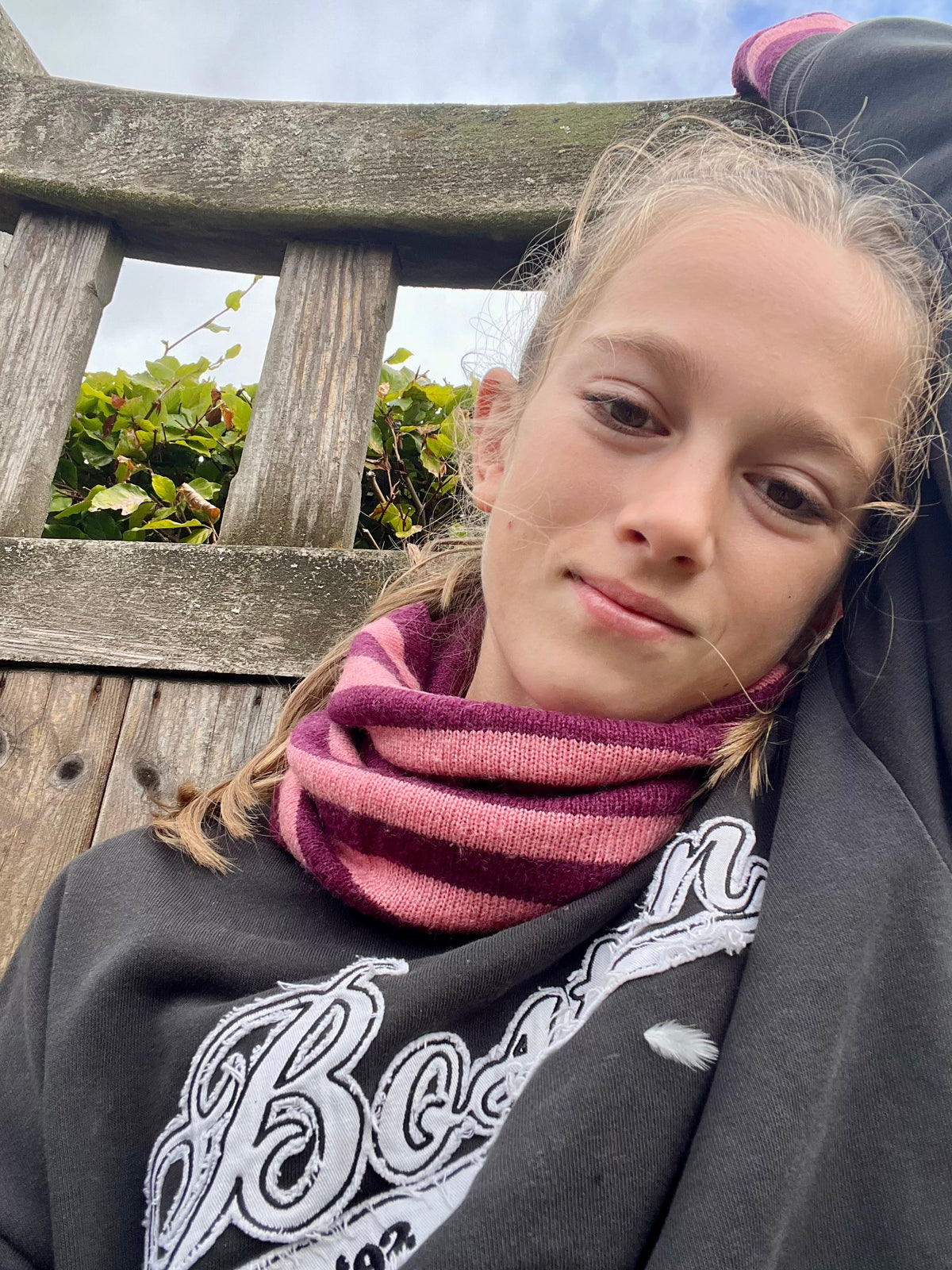 A close-up lifestyle shot of a young woman outdoors leaning against a weathered wooden bench. She is wearing a dark gray sweatshirt with a white graphic print and a thick, striped cashmere snood in shades of pink and burgundy.