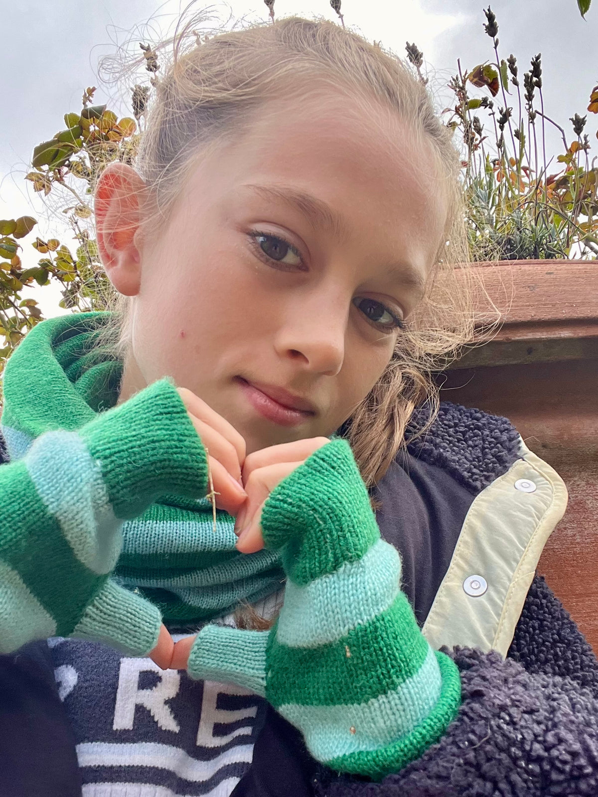 A close-up lifestyle shot of a young girl wearing green and light blue striped knit fingerless cashmere gloves and a matching green knit snood, set against an outdoor garden background.