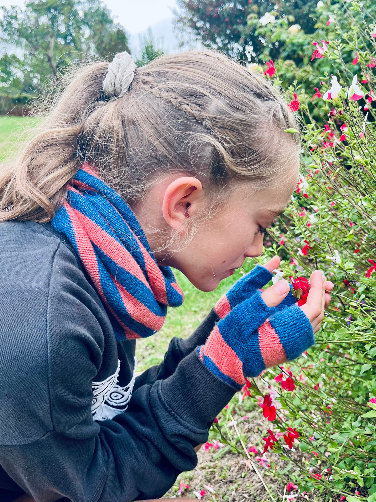 A child outdoors wearing a blue and pink striped knit cashmere snood and matching fingerless gloves, looking down at red flowers in a garden.