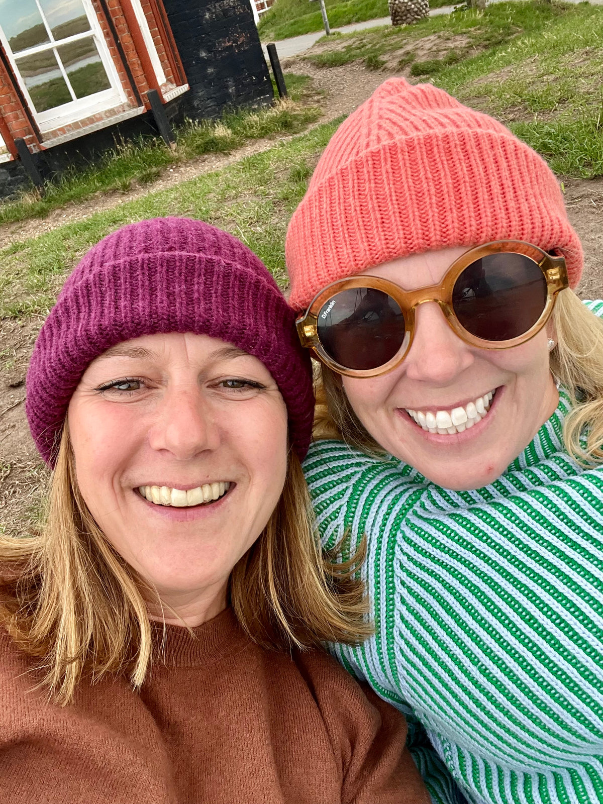 Two women smiling for a selfie outdoors, each wearing a chunky knit cashmere beanie. The woman on the left wears a burgundy beanie and a brown top, while the woman on the right wears a coral beanie, round brown sunglasses, and a green and white striped sweater.