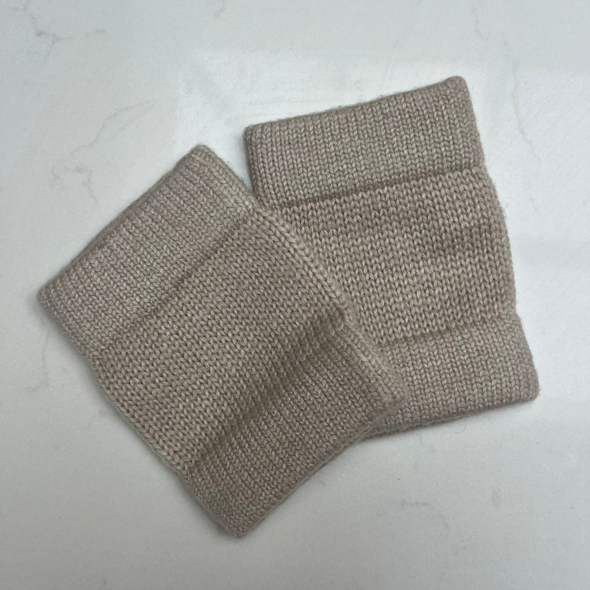 A pair of oatmeal cashmere knit cuffs to wear on your wrists with ribbed elastic edges, displayed on a light gray marble-textured background.