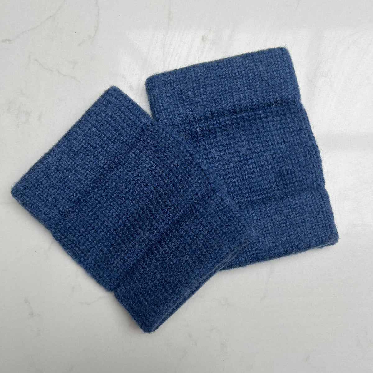 Two soft navy blue knit cashmere cuffs to wear on your wrists with ribbed elastic edges, displayed on a light gray marble-textured background.