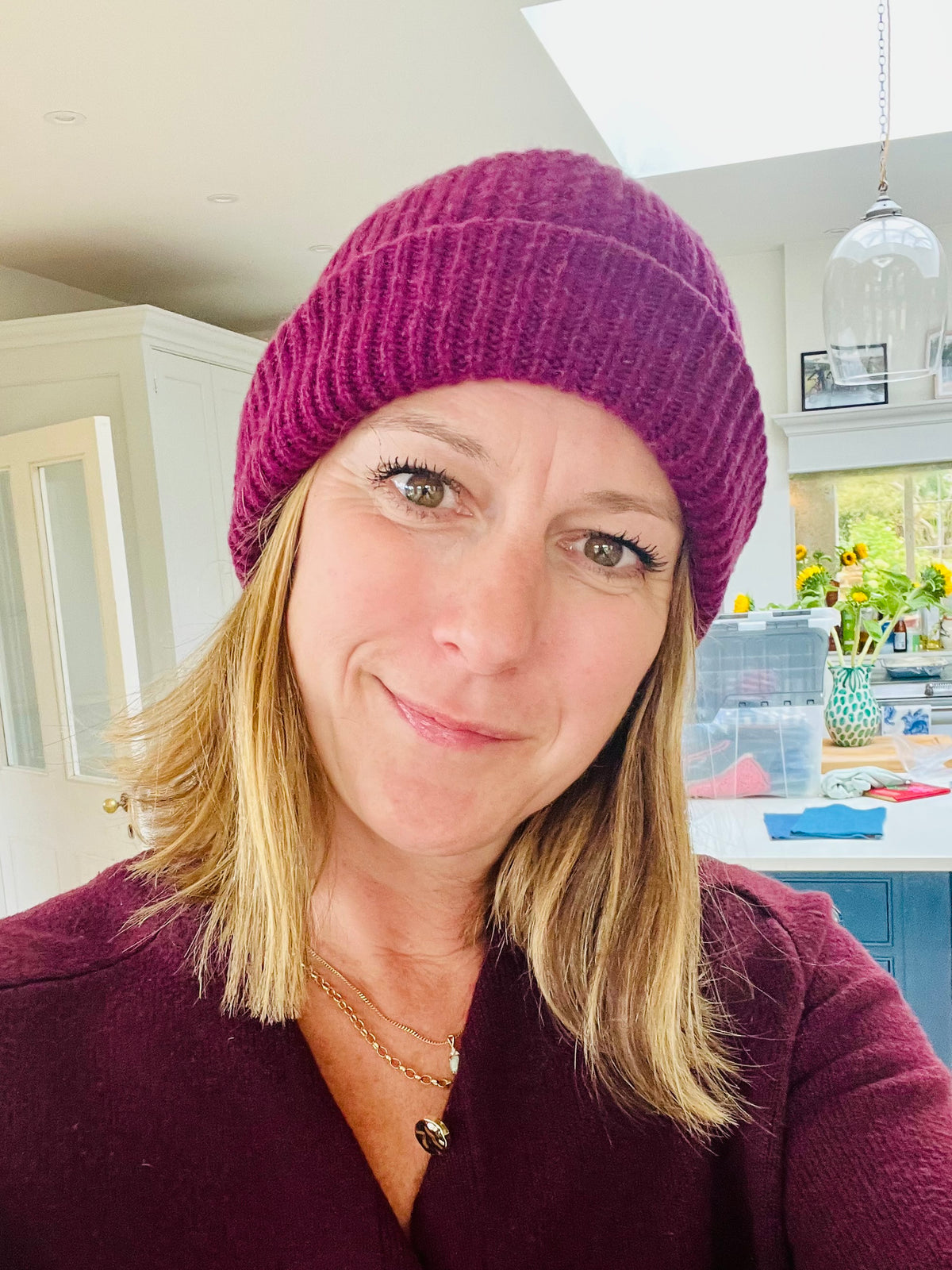 A woman wearing a ribbed knit cashmere beanie in a deep burgundy colour, photographed in a bright kitchen setting.