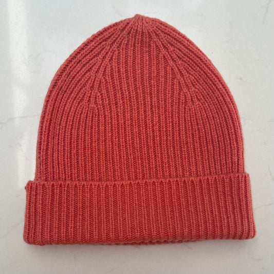 A single ribbed knit beanie hat in a bright coral color, laid flat on a light gray marble surface.