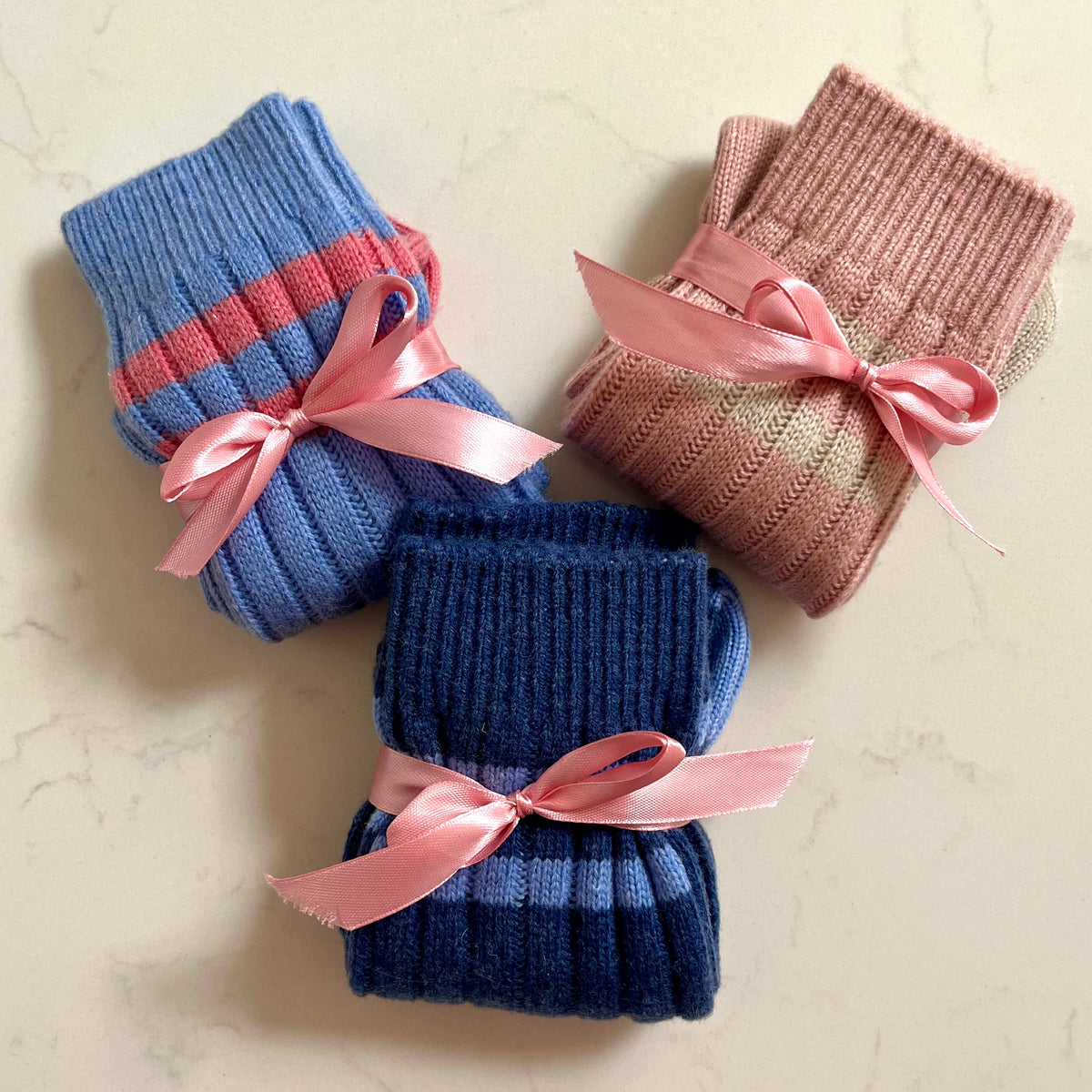Three pairs of folded ribbed knit cashmere socks tied with pink satin ribbons, arranged on a light beige marble surface. One pair is light blue with two pink stripes near the top, one is dusty pink with two beige stripes, and one is dark navy blue with two light blue stripes.