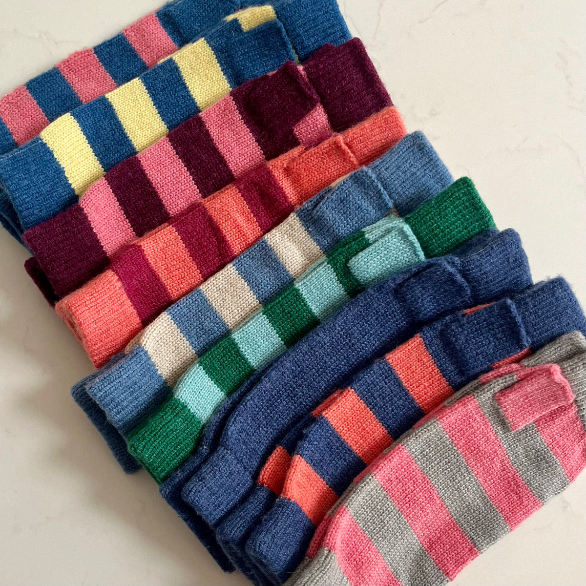A stack of folded striped knit cashmere wristwarmers in multiple colours, including blue, pink, yellow, green, orange, and gray, arranged diagonally on a light grey marble surface.