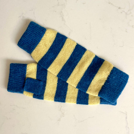 A pair of knitted cashmere wristwarmers with alternating royal blue and pale yellow stripes, laid flat on a light marble surface.