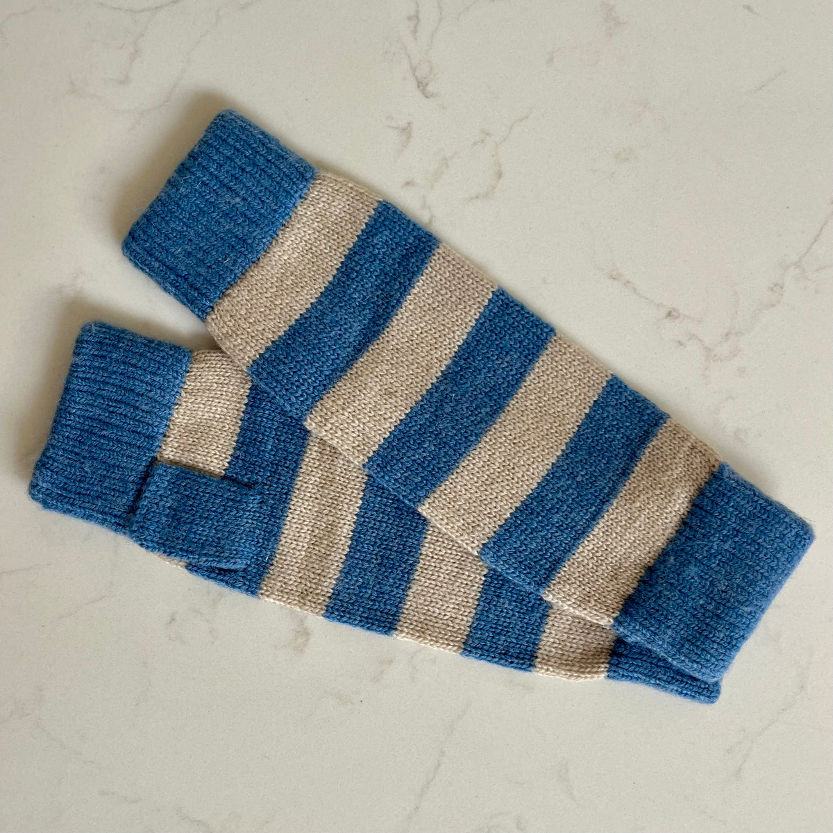 A pair of blue and beige striped knit cashmere wristwarmers laid flat on a light marble surface.