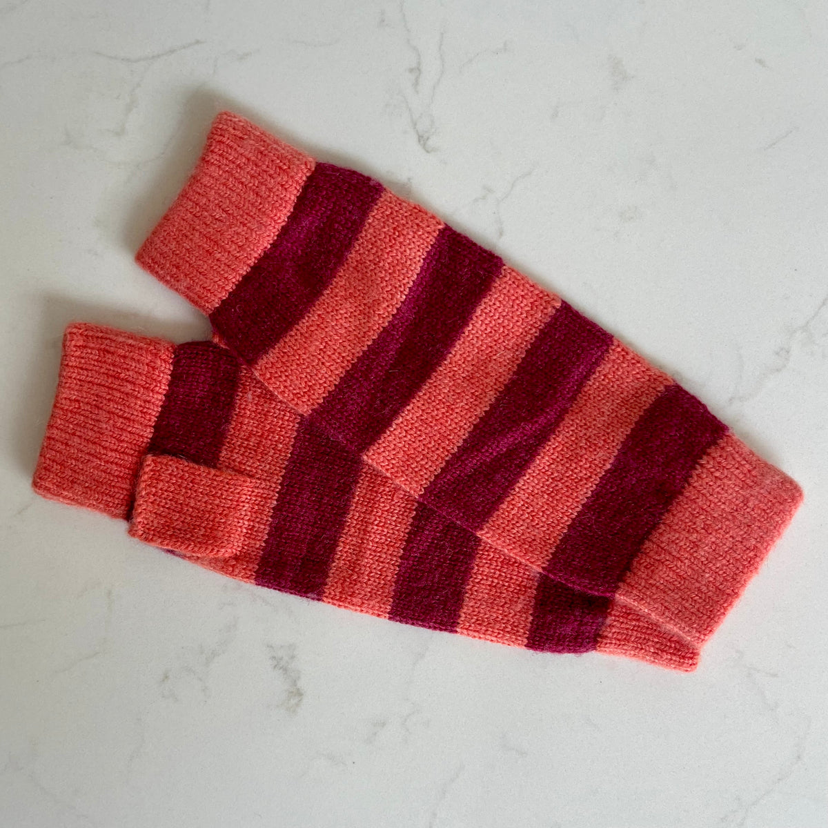 A pair of knit fingerless cashmere gloves with horizontal stripes in hot coral orange and deep burgundy pink, laid flat on a light gray marble surface.