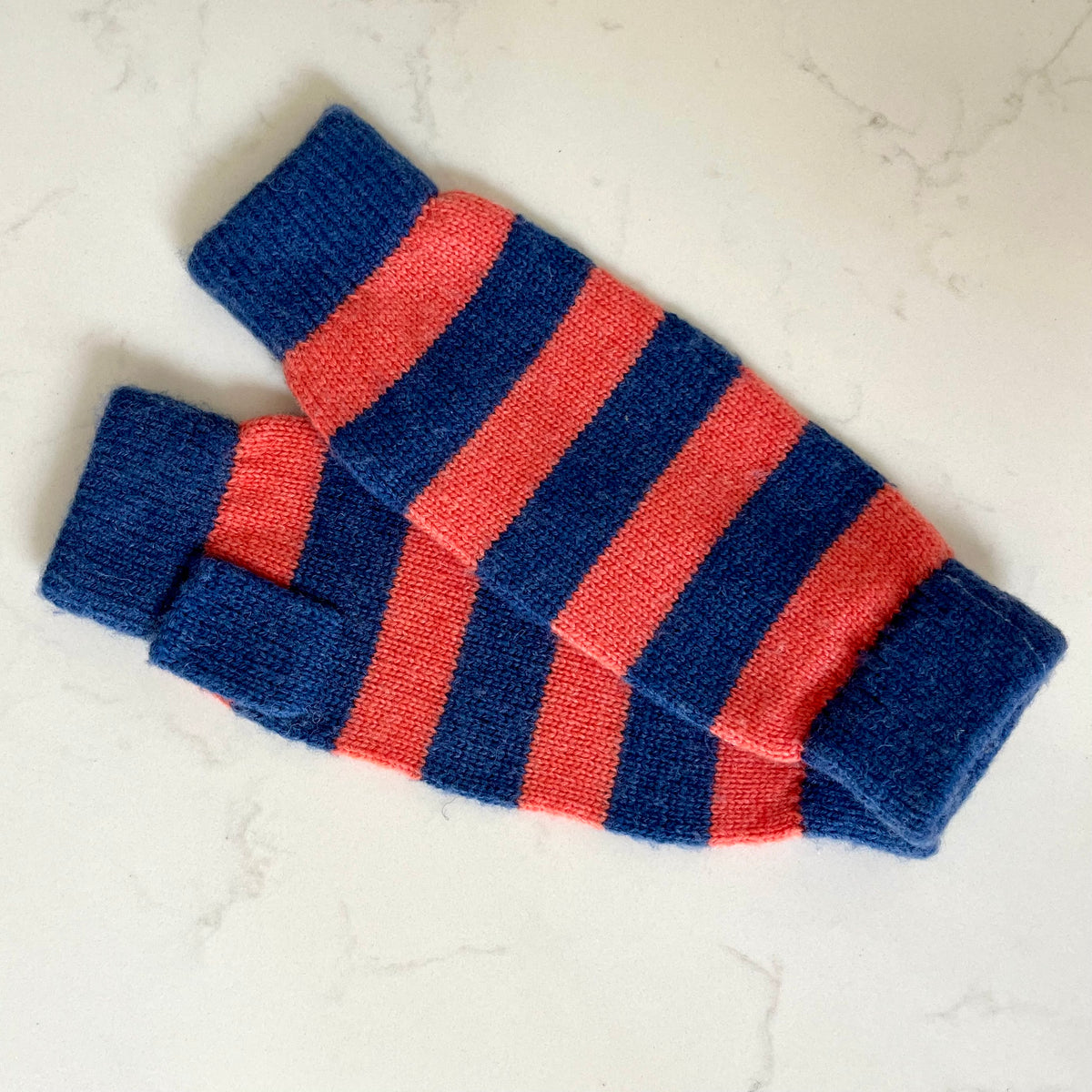 A pair of knitted cashmere fingerless gloves with alternating navy blue and coral stripes, laid flat on a light marble surface.
