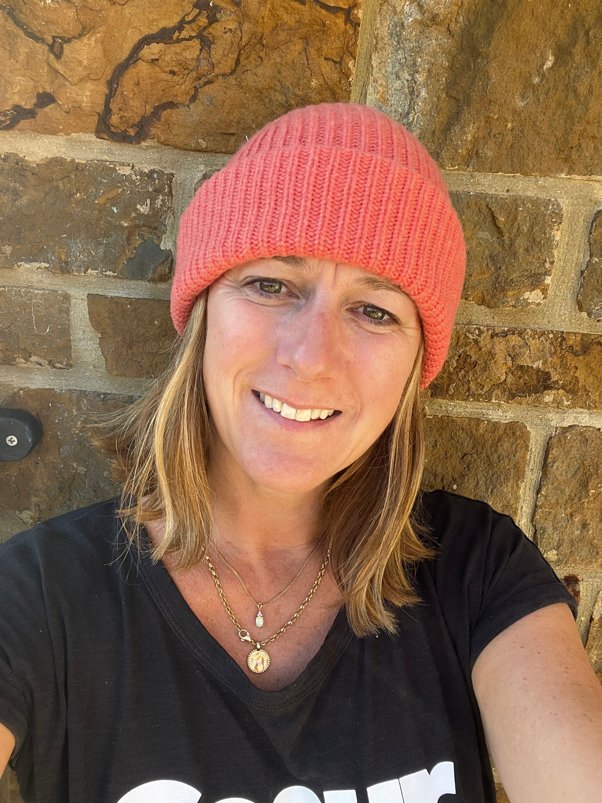 A smiling woman wearing a coral pink ribbed knit cashmere beanie hat and a black t-shirt, posing in front of a rustic stone wall.