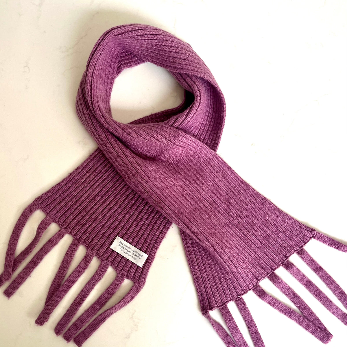 A lilac coloured, ribbed knit cashmere mix kids scarf, featuring fringed ends and a small white brand label, laid flat on a light off-white background.