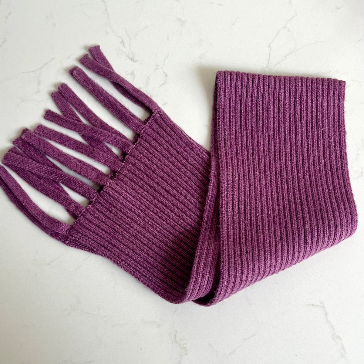 A lilac purple ribbed knit scarf with fringe tassels, laid flat on a light gray marble surface.