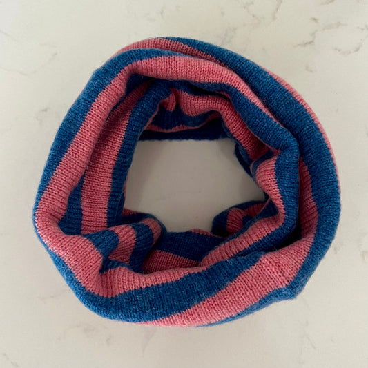 A knitted kids cashmere snood with alternating horizontal stripes of dark blue and pink, displayed on a light marble surface.
