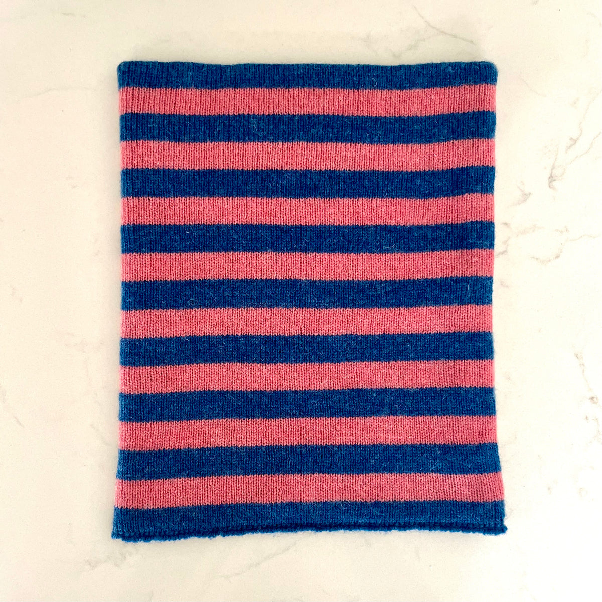 A rectangular knit fabric cashmere snood featuring horizontal stripes in navy blue and pink, laid flat on a light marble surface.