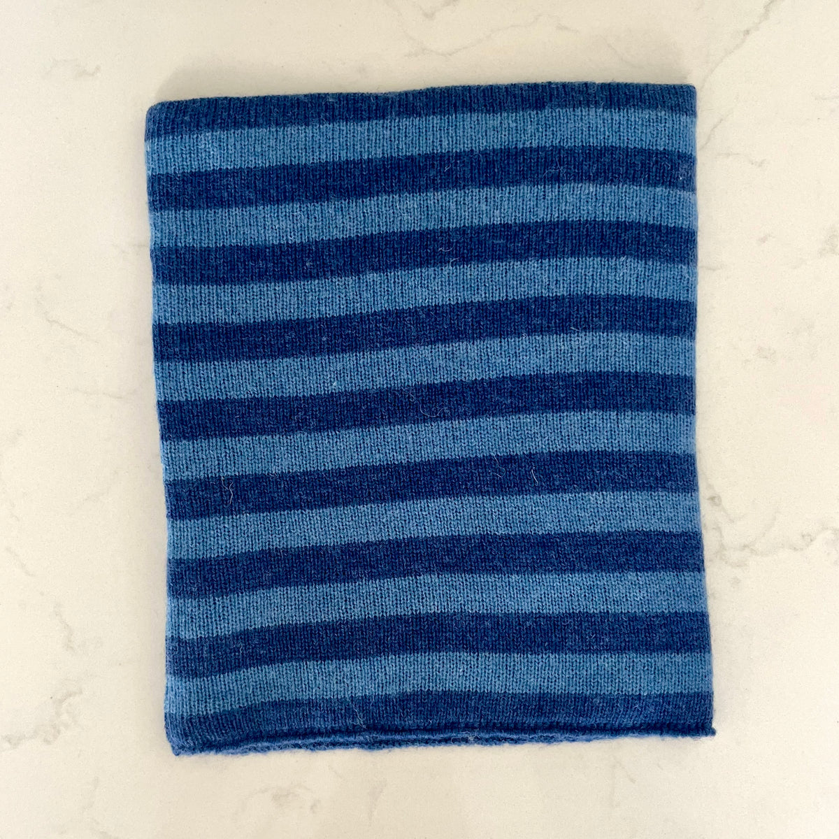 A kids knitted cashmere snood, featuring horizontal stripes in shades of blue and navy, laid flat and displayed on a light marble surface.