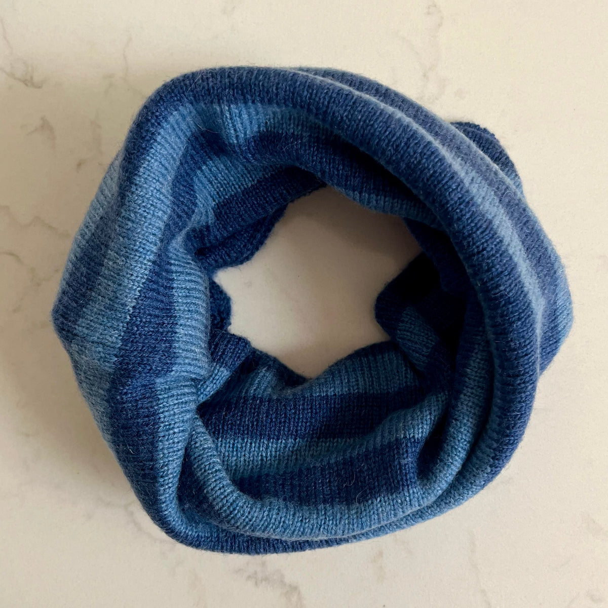 A blue striped knitted kids cashmere snood, folded into a loop, displayed on a light beige marble surface.