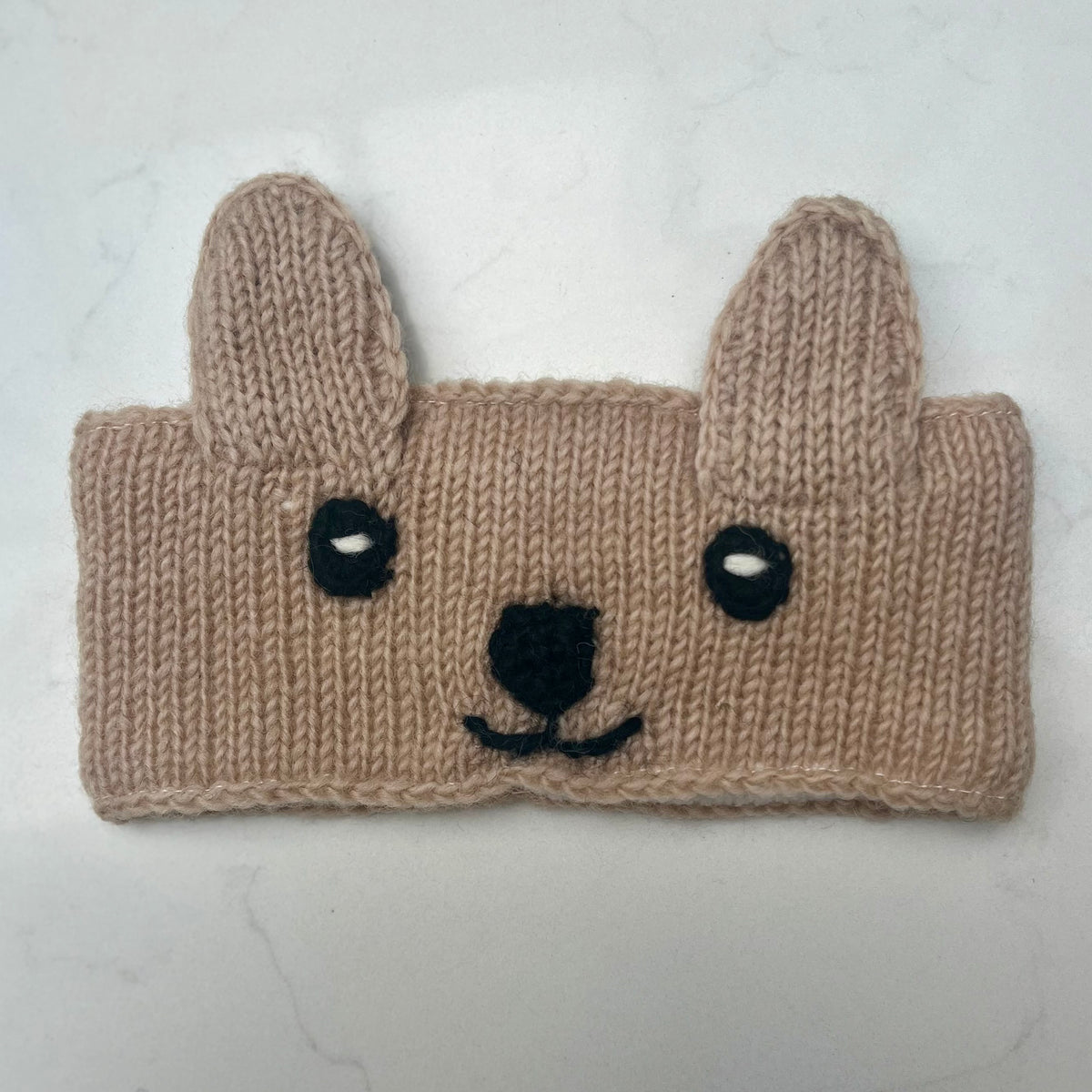 A handmade knitted cream headband designed to look like a cute bears face, featuring floppy ears, black embroidered eyes, a black nose, and a small black smile, shown on a light marble background.