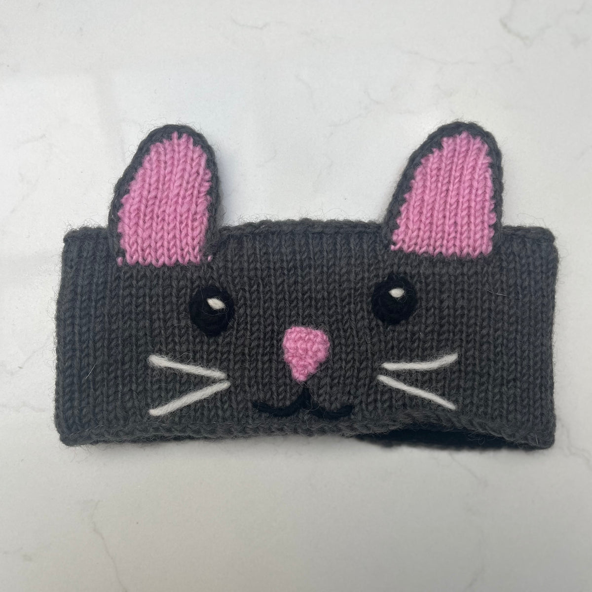 A dark gray knitted cat face headband with pink ears, a pink nose, black eyes, and white whiskers, shown on a light marble background.