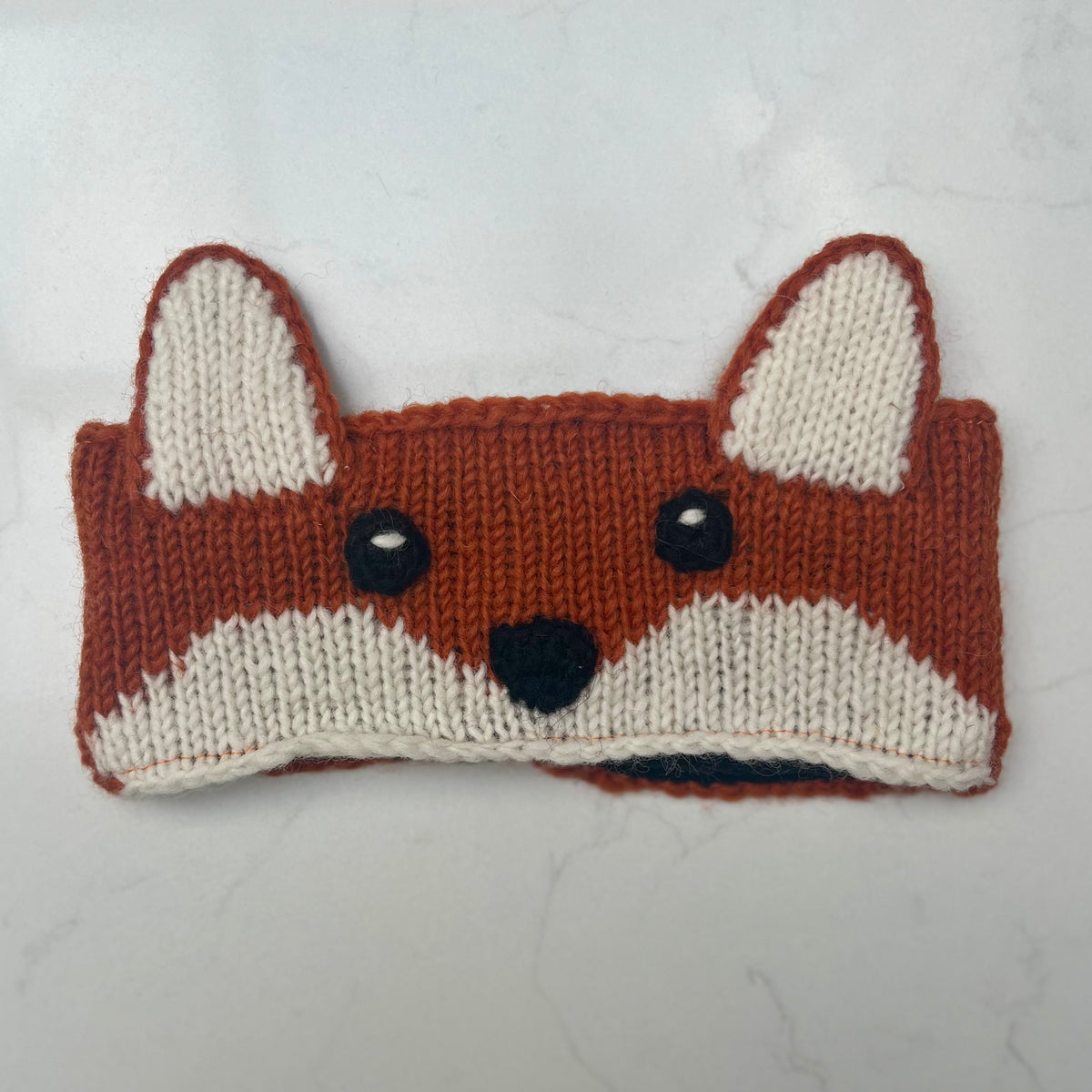 A handmade knitted headband shaped like a fox face, featuring a rust-orange and cream knit pattern with black yarn eyes and nose, displayed on a light gray marble surface.