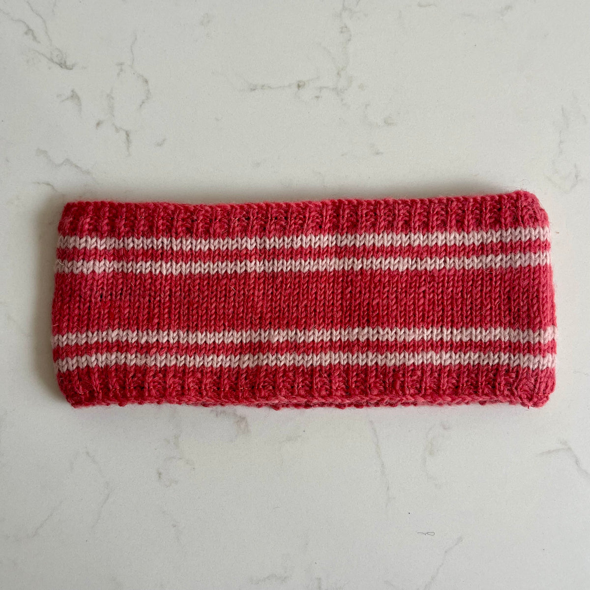A coral knit headband with thin white horizontal stripes, laid flat on a light gray marble surface.