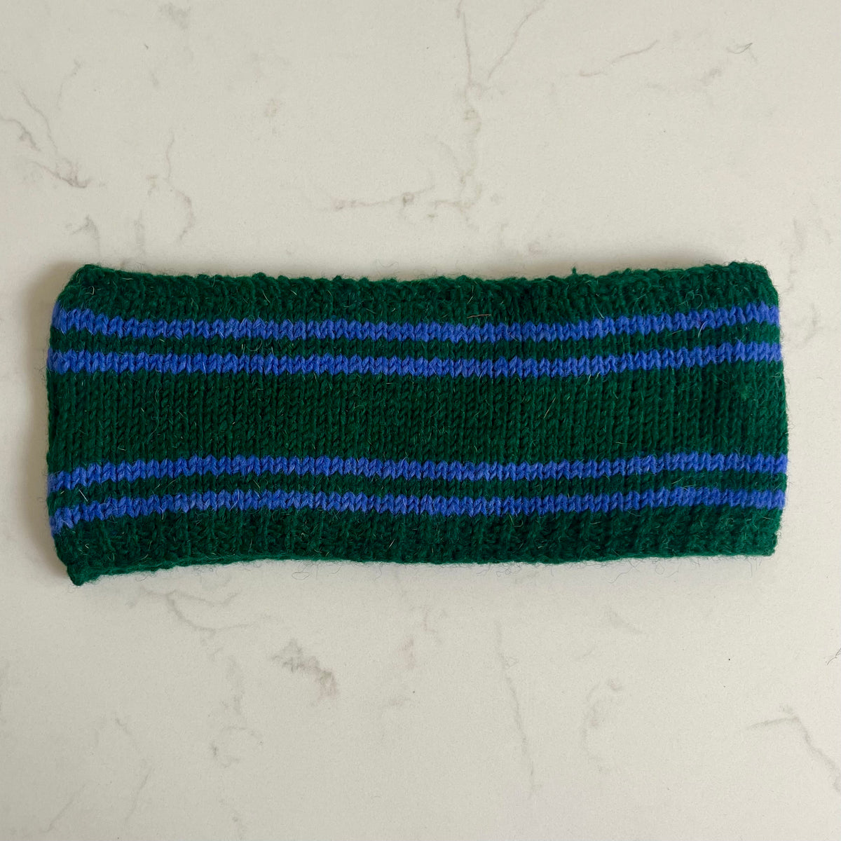 A dark green knitted headband with two horizontal light blue stripes, laid flat on a light gray marble surface.