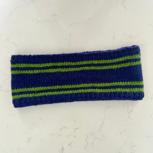 A handmade knit headband in navy blue with lime green horizontal stripes, laid flat on a light gray marble surface.