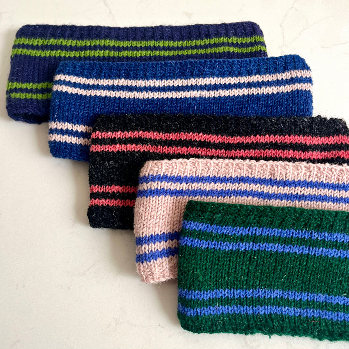 Five knitted wristbands with horizontal stripes in various colour combinations, arranged diagonally on a light off-white background.