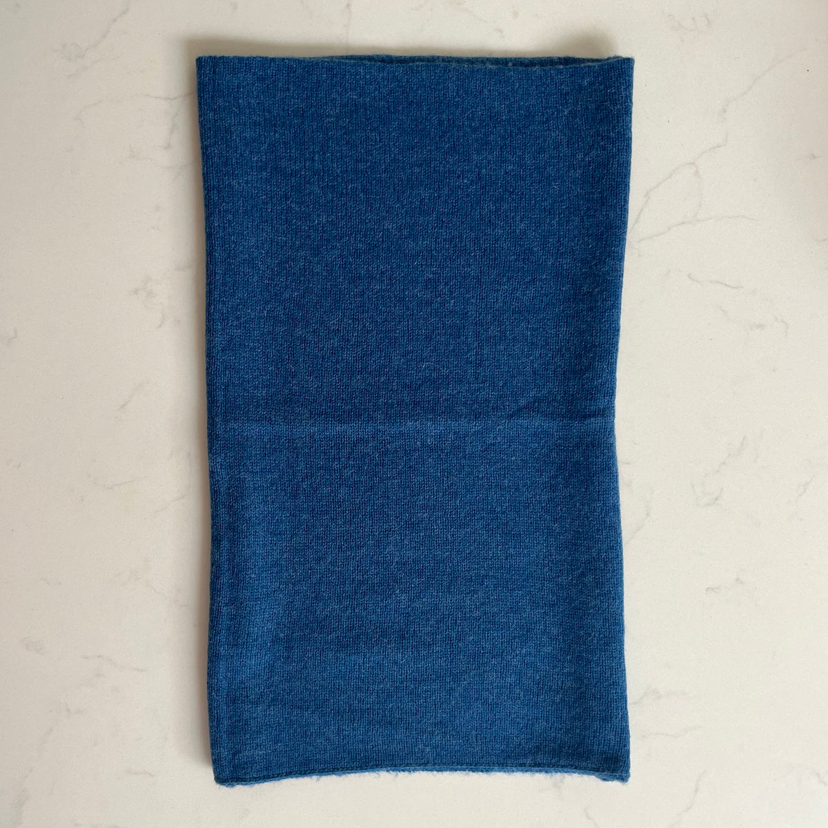 A folded dark blue knit cashmere snood item, laid flat on a light beige marble surface.