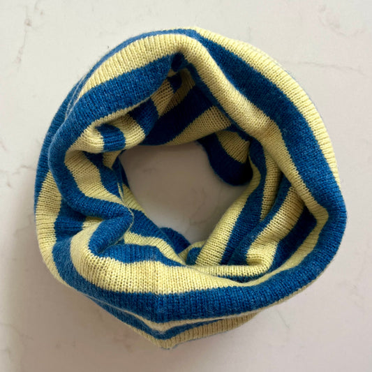 A knitted cashmere snood with alternating blue and pale yellow stripes, displayed in a loop on a light marble surface.