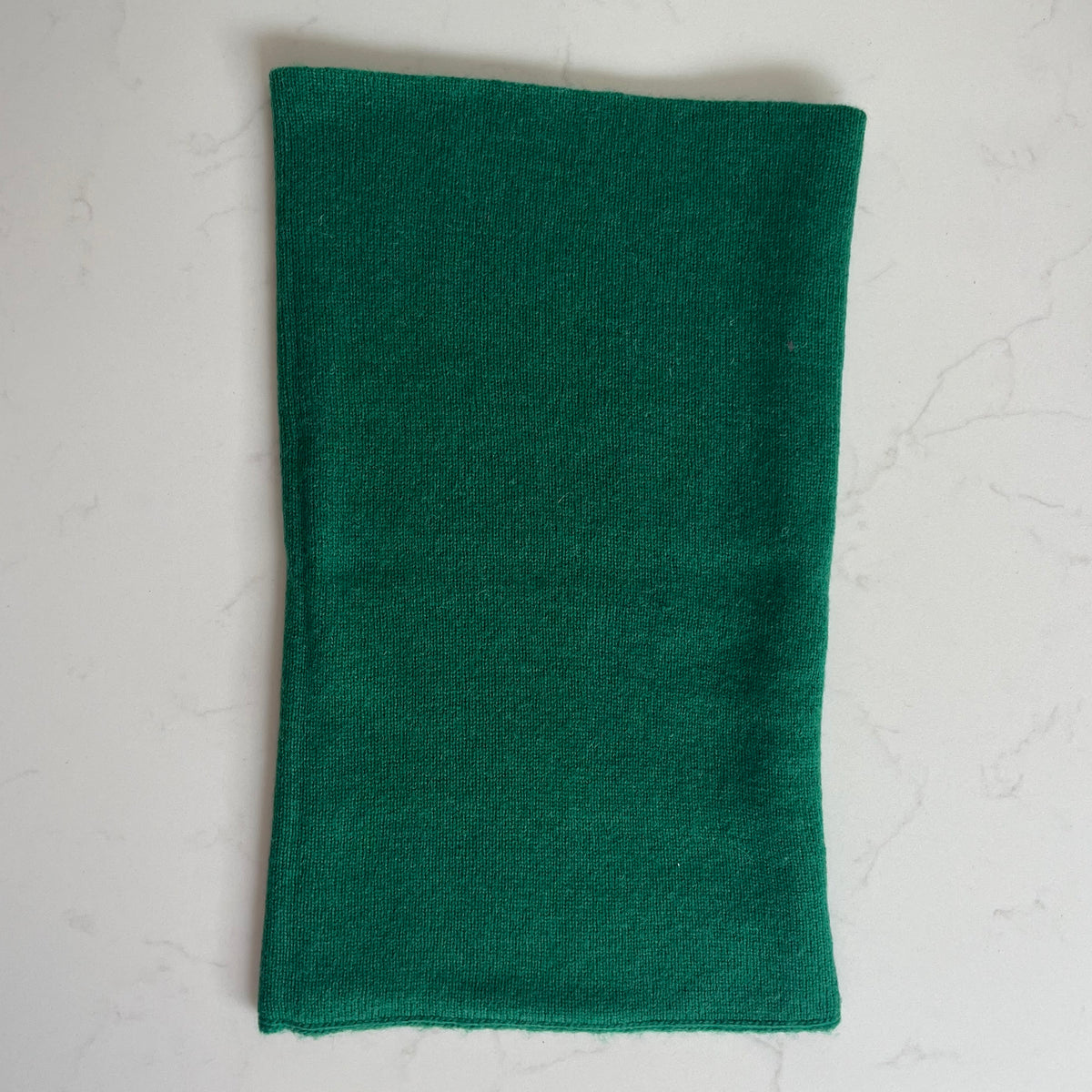 A dark green cashmere snood displayed flat on a light gray marble surface.