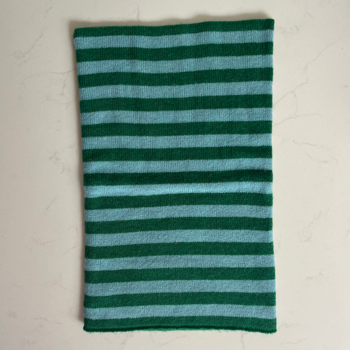 A rectangular striped knit cashmere snood featuring alternating horizontal stripes of dark forest green and light blue, laid flat on a light beige marble surface.