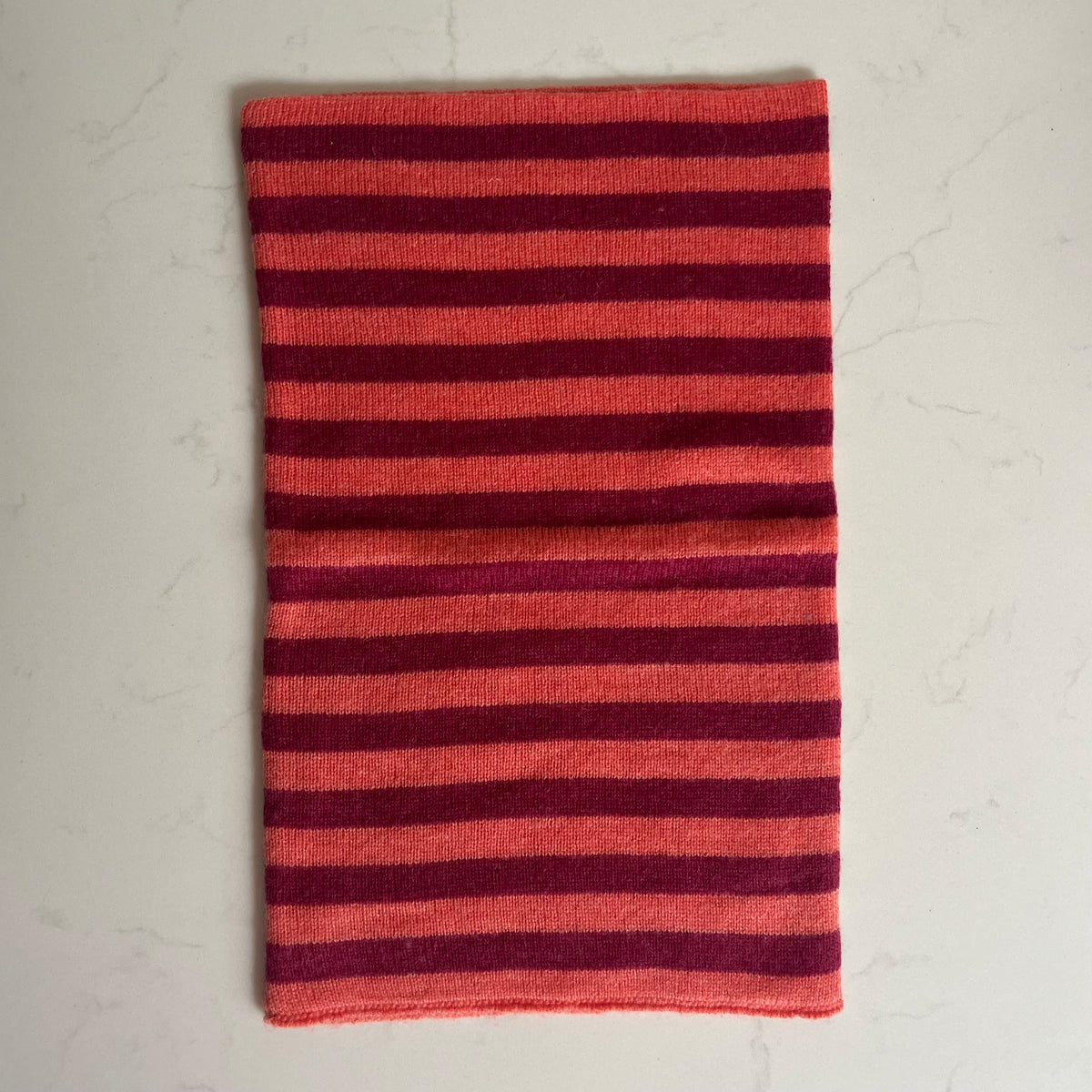 A rectangular striped knit cashmere snood, featuring horizontal stripes in hot coral and raspberry pink, laid flat on a light marble surface.