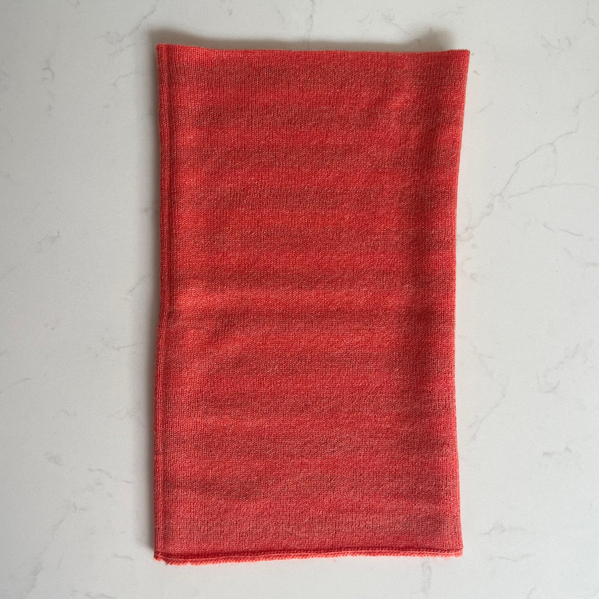A folded hot coral coloured cashmere snood, laid flat on a light gray marble surface.