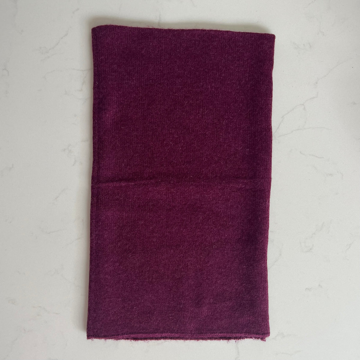 A rectangular cashmere burgundy snood, laid flat on a light gray marble surface.