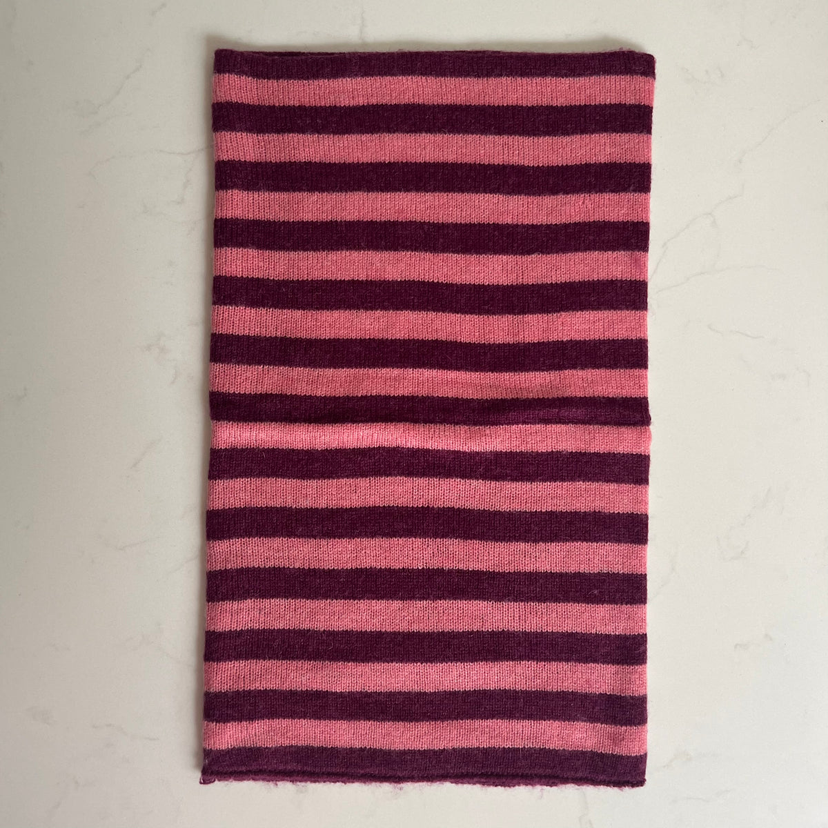 A rectangular striped knit cashmere snood, featuring alternating horizontal stripes of dark burgundy and soft pink, laid flat on a light marble surface.