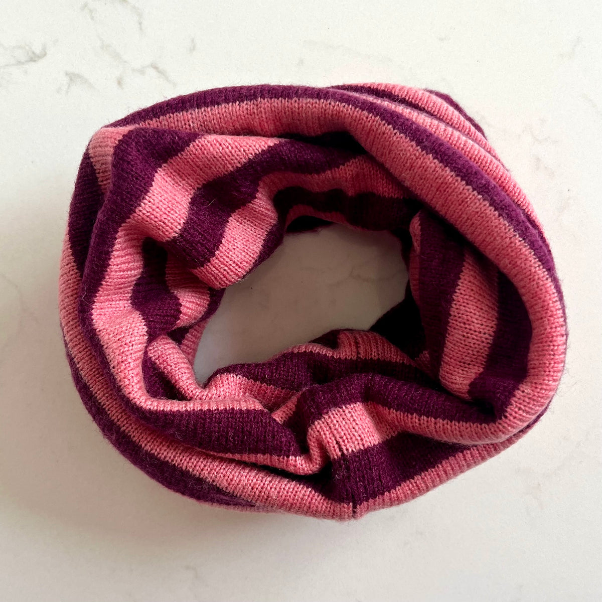 A burgundy & pink cashmere snood, photographed flat on a light marble-textured background.