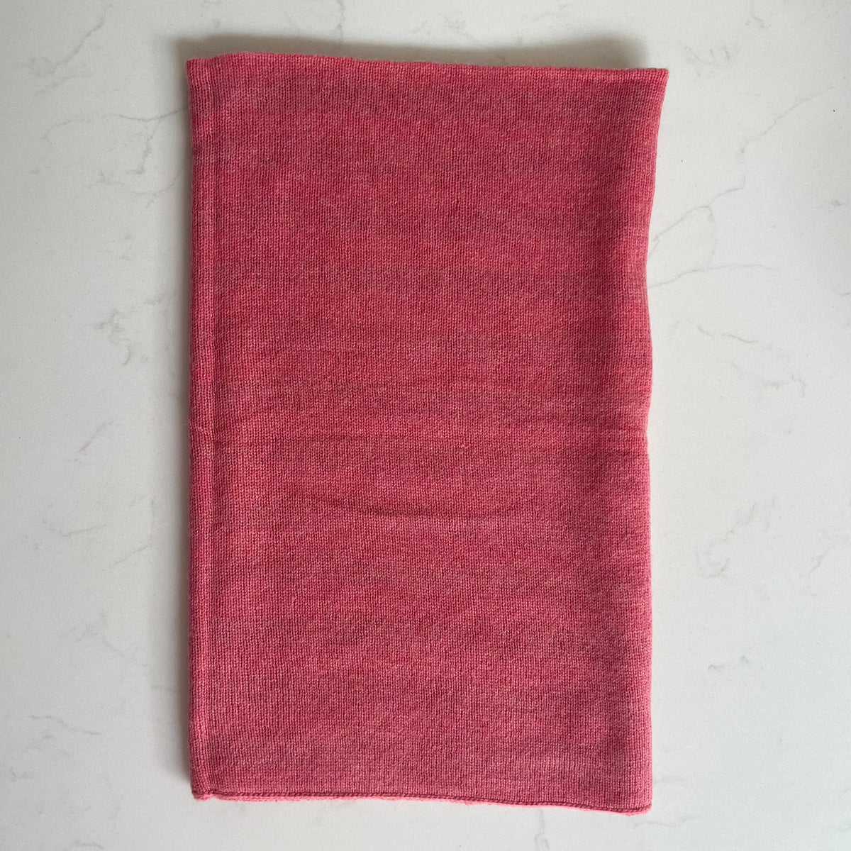 A folded pink cashmere snood, laid flat on a light gray marble background.
