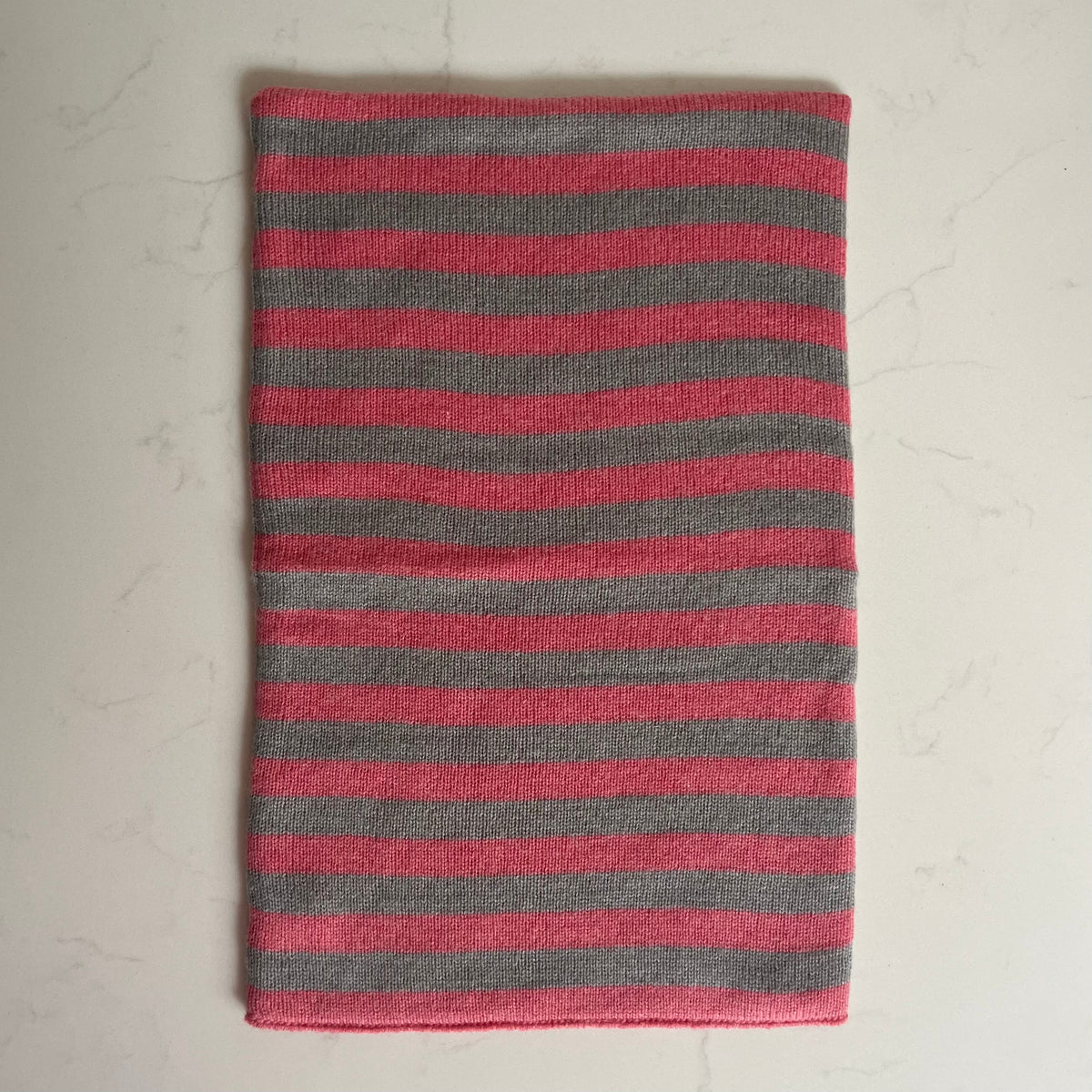 A rectangular striped cashmere snood with alternating pink and gray horizontal stripes, laid flat on a light marble-like surface.