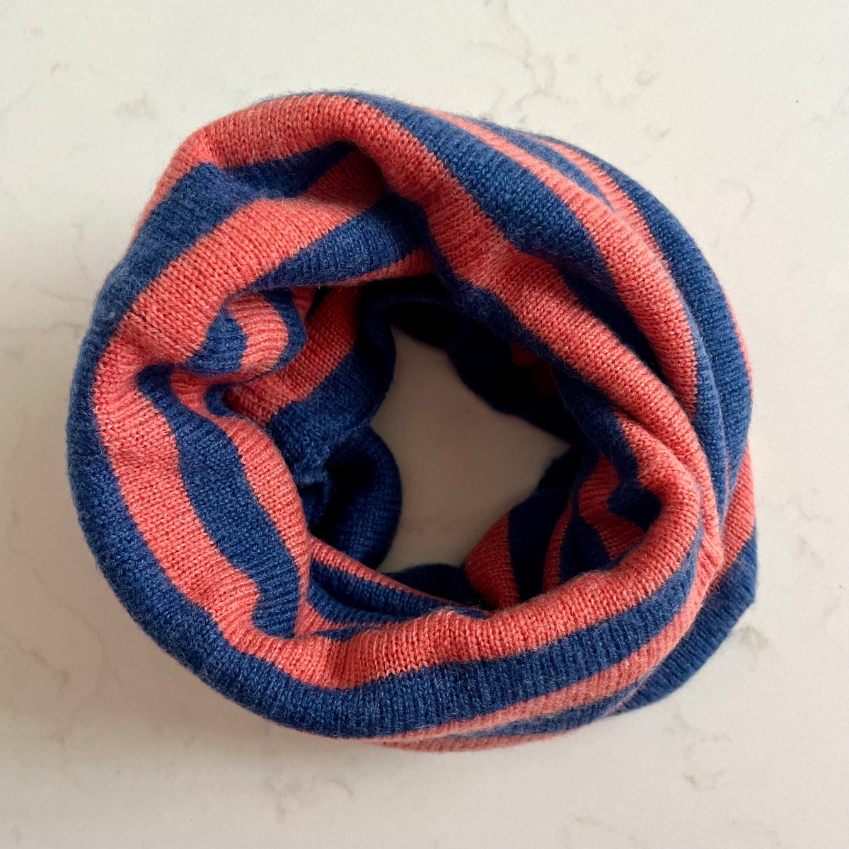 A striped knit cashmere snood with alternating soft navy blue and coral pink stripes, displayed on a light marble surface.