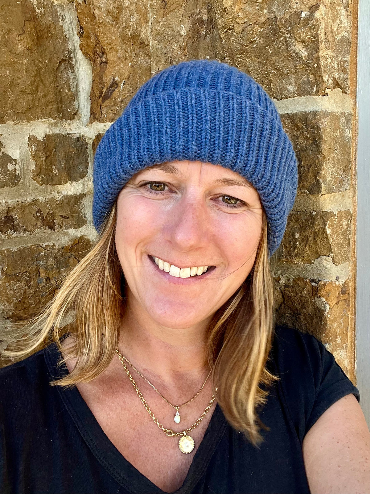 A smiling woman with blonde hair wearing a soft navy blue ribbed knit cashmere beanie hat and a black top, standing in front of a rustic stone wall.