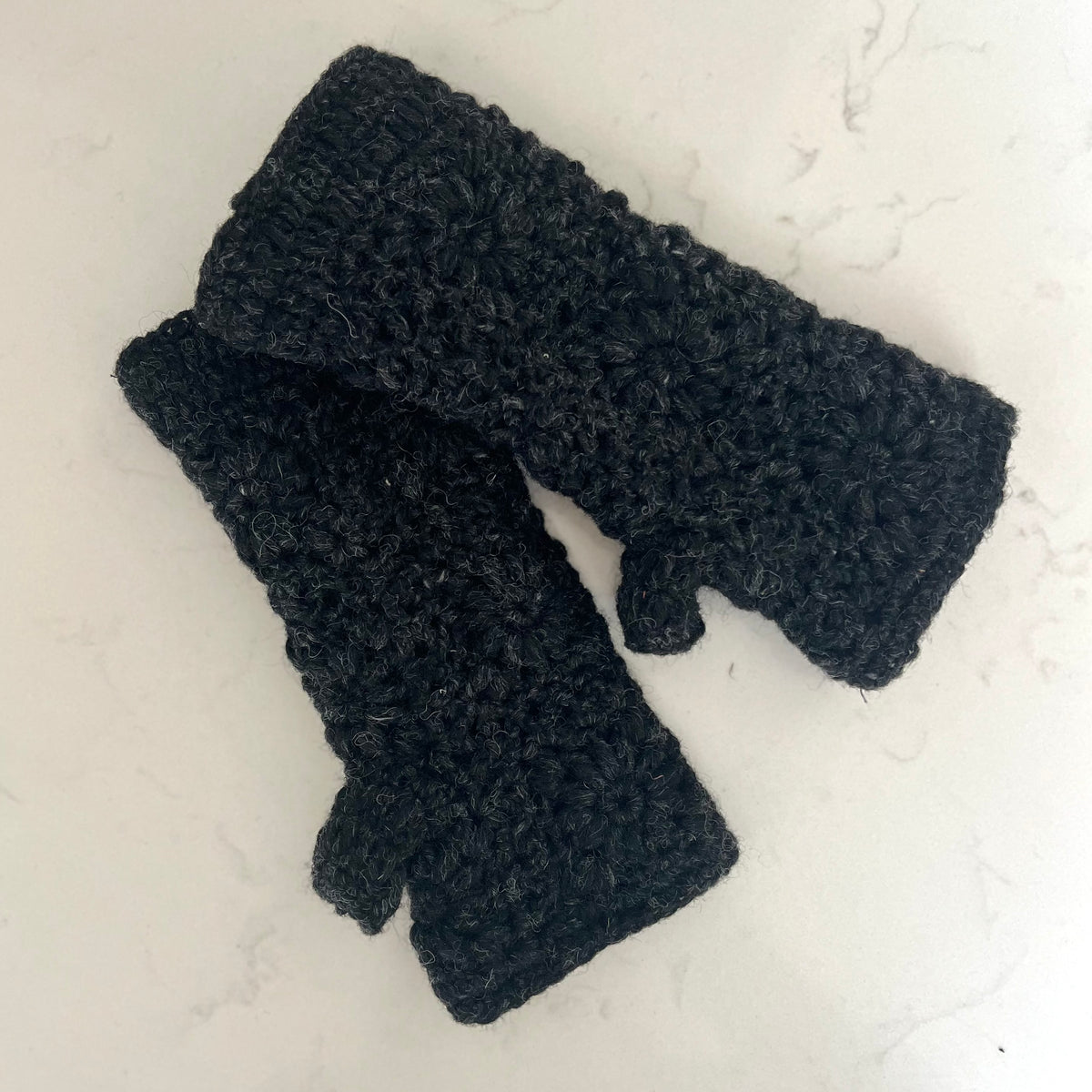 A pair of chunky charcoal crocheted wristwarmers laid flat on a light off-white marble surface.