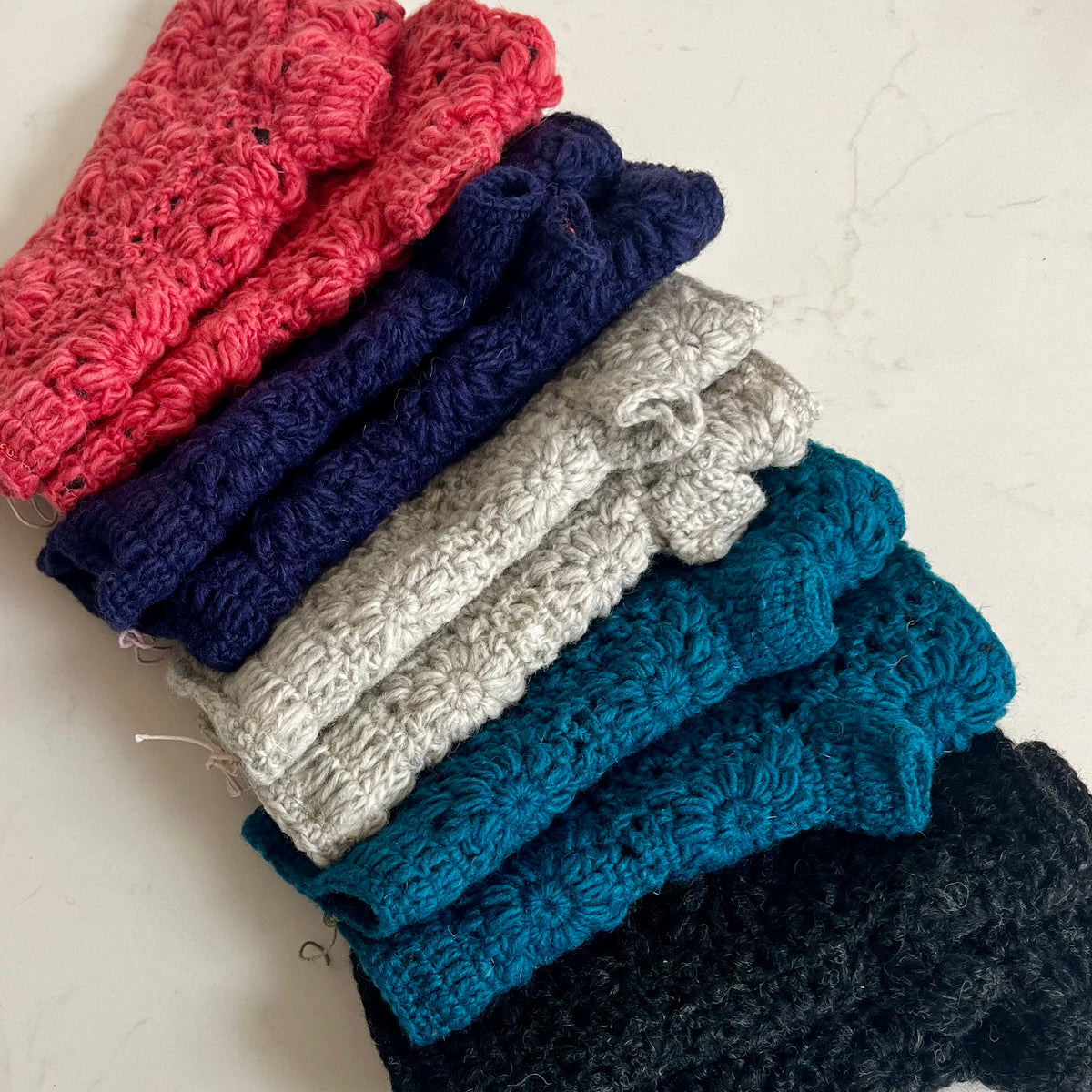 A stack of textured crochet knit wristwarmers in multiple colors, including pink, navy, light gray, teal, and black, arranged diagonally on a light marble surface.
