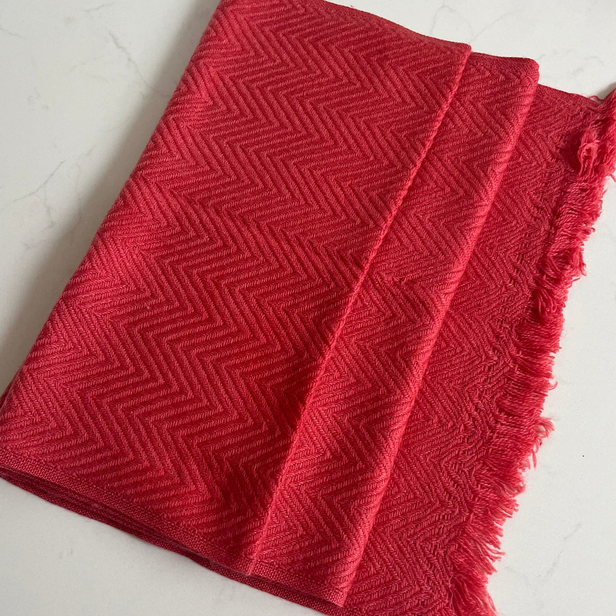A folded coral red woven cashmere scarf with a textured chevron pattern and fringed edges, photographed on a light marble-like surface.