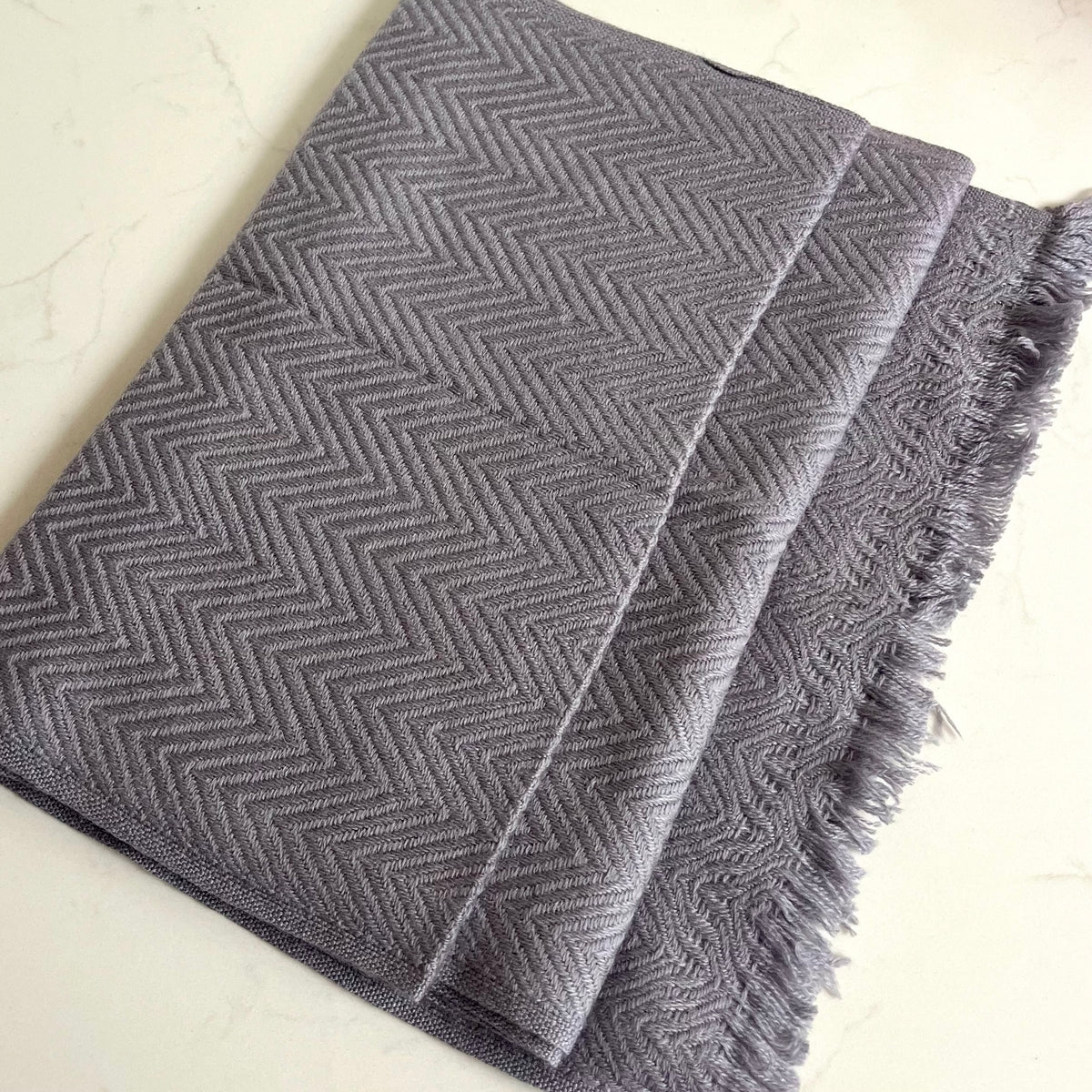 A folded gray woven cashmere scarf with a herringbone pattern and fringed edges, photographed on a light marble-like surface.