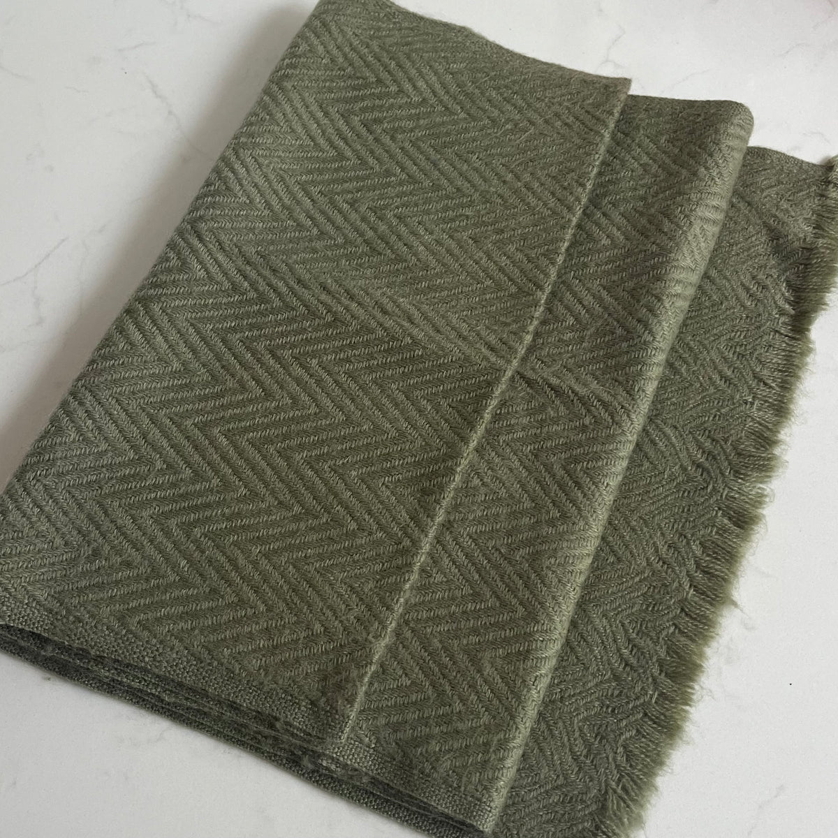 A folded olive green woven cashmere scarf with a herringbone pattern and fringed edges, displayed on a light marble surface.